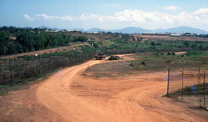 18. The Phu Cat AB perimeter on the east side of the base, from Tango 4 looking south. On the horizon: the ROK Army camp on the left, the base barracks on the right. From L to R: the Village adjacent to the base, the railroad, the physical perimeter (barbed wire, tanglefoot, belts of concertina, lights). The trip flares in the concertina are not visible. Supply yard fence to the right. Photo by Don Bishop. 1969-1970.